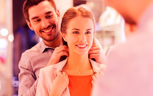 Imagem representando a Experiência do cliente, contendo uma mulher experimentando um colar, com a ajuda de um homem em uma loja física.